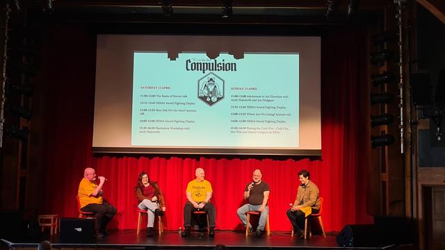 Theatre stage, wood surround and beams overhead, red drapes backdrop, projector screen in front with black amd white Conpulsion logo of owl style mage rolling dice, list of days sessions left and right on stage, 5 seated panellists talking on microphones