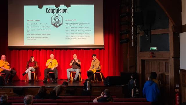 Theatre stage, wood surround and beams overhead, red drapes backdrop, projector screen in front with black amd white Conpulsion logo of owl style mage rolling dice, list of days sessions left and right on stage, 5 seated panellists talking on microphones