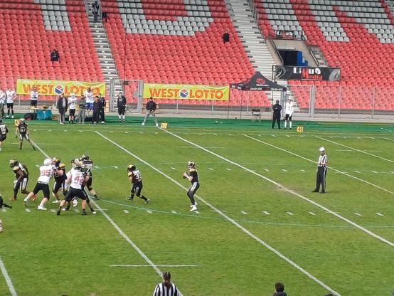 Das Bild zeigt eine American-Football-Spielzeit auf einem grünen Rasenfeld. Im Vordergrund sind Spieler in schwarzen und weißen Trikots zu sehen, die sich in einer Offensiveformation befinden. Ein Quarterback in schwarzer Uniform hält das Ball und scheint sich auf einen Wurf vorzubereiten. Die Spieler tragen Helme und Schutzausrüstung. Ein Referee in schwarz-weiß gestreifter Uniform steht auf dem Spielfeld und beobachtet die Spielverlauf. Im Hintergrund sind rote und graue Stadiontribünen zu sehen, die teilweise mit Zuschauern besetzt sind. An den Tribünen hängen Banner mit der Aufschrift "WEST LOTTO" und "Liux Media". Die Spielfeldmarkierungen sind deutlich sichtbar, und die Linienlaufzahlen 40 und 50 sind markiert.

Bereitgestellt von @altbot,