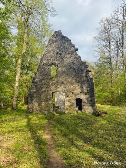 Frontansicht der Ruine der Kirche St. Valentin bei Laubach. Das spitz zulaufende Giebeldach aus grobem Naturstein steht noch erhalten. Ein schmaler Trampelpfad führt durch das Portal in das Innere der ehemaligen Kirche. Die Ruine ist von frischem Frühlingsgrün und hohen Bäumen umgeben.