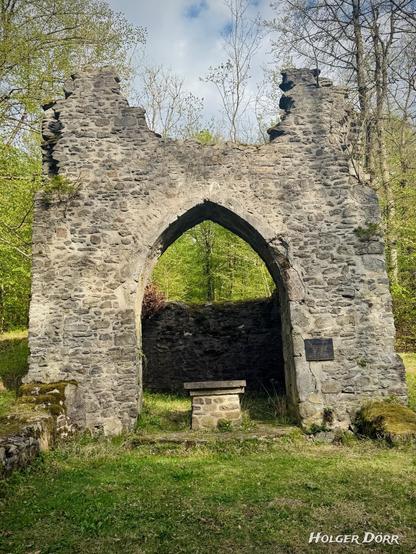 Blick durch den gotischen Spitzbogen im Inneren der Ruine. Vor einer halb erhaltenen Rückwand steht ein schlichter Altar aus Bruchstein. Die Umgebung ist grün und lichtdurchflutet. Rechts am Mauerwerk ist eine dunkle Metalltafel angebracht.
