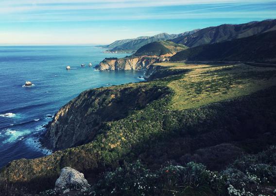 The blue Pacific Ocean is in the left of the frame. Along the center is a jagged coastline. To the right, green hills and mountains beyond.