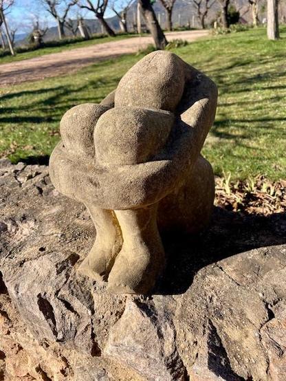 A photo of a sculpture in stone of a huddled human being, sitting with the arms around the knees. Artist unknown. The sculpture is placed on a lower stone wall. It’s outdoor art, the sculpture is situated in a natural park-like setting with grass and trees. 