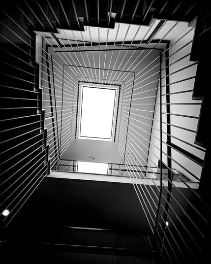 Ein Treppenhaus in schwarz-weiß von unten fotografiert, in der Mitte das Oberlicht, rundherum die Treppenstufen, weitgehend versteckt hinter den Treppengeländer-Stangen, die gleichmäßig verteilt links, oben und rechts um das Bild und auf das Oberlicht zu laufen.