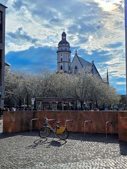 Dieses Foto wurde in Leipzig aufgenommen und zeigt einen malerischen Blick auf die Thomaskirche und die Thomaswiese.
Im Vordergrund befindet sich ein gepflasterter Platz mit einem Fahrradständer, an dem ein Fahrrad abgestellt ist. Der Boden ist mit kleinen, unregelmäßigen Pflastersteinen bedeckt, die eine leicht holprige Oberfläche erzeugen.
Im Hintergrund erhebt sich die imposante Thomaskirche mit ihrem hohen Turm und spitzen Dach. Die Kirche ist von blühenden Zierkirschen umgeben, die mit ihren weißen Blüten einen schönen Kontrast zum blauen Himmel bilden. Die Zierkirschen blühen auf der Thomaswiese, einem öffentlichen Platz in Leipzig.
Der Himmel ist von hellen, flauschigen Wolken bedeckt, die sich über die Szene erstrecken. Das Sonnenlicht scheint von oben und erzeugt einen hellen Fleck in der oberen rechten Ecke des Bildes.
In der Mitte des Bildes befindet sich ein kleines Gebäude mit der Aufschrift "Curry CULT". Es scheint sich um einen Imbissstand oder ein kleines Restaurant zu handeln.
Am unteren Bildrand befindet sich ein hellblauer Schriftzug mit der Aufschrift: "F.loki ~ Enjoy the moment".