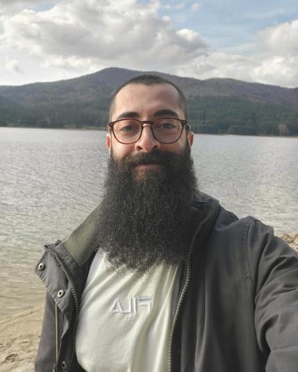 Me, a bearded young man with glasses standing with a a lake and mountains behind me