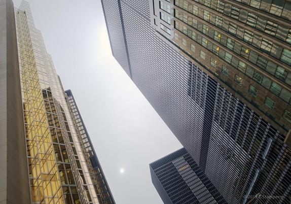 Looking up at skyscrapers.  The one to the left has its left edge aligned vertically and the rest of the buildings converge towards that line of perspective, giving a disorienting feeling.  Between the buildings we can see a dim light of the sun trying to break through clouds. 