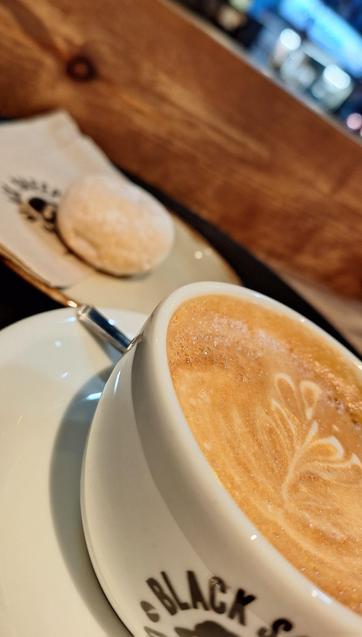 Angled photo of a coffee cup with latte art, part of the print "Black S" is visible at the front. in the background a small sugar-coated pastry, a wooden panel, and a dark street with shop lights are visible.