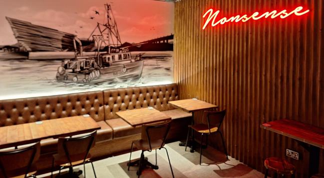 Picture of a corner of a cafe with chairs and a bench on the wall. There is a mural in black and white on the left corner wall, taking up most of the photo, a ship is in the Tay with the V&A in the background. The right side wall has some wooden panelling and a neon sign in handwritten style saying "Nonsense".