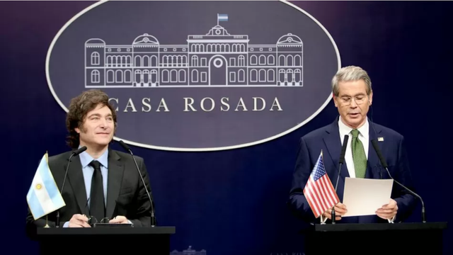 Imagen de dos hombres con corbata y traje. A la derecha el Secretario de Estado de EEUU leyendo un papel. Tiene una bandera de su país junto al micrófono. A la izquierda, y más abajo en el plano, el presidente argentino sonríe escuchando atento el discurso del Secretario. De fondo, un cartel dice "Casa Rosada". El fondo es negro y los hombres resaltan sobre el fondo.