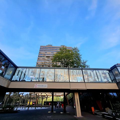 Een glazen loopbrug met daarop in grote witte letters de tekst "DOE IETS WAT ERTOE DOET". Op de achtergrond is het Erasmusgebouw van de Radboud Universiteit in Nijmegen te zien, met een heldere blauwe lucht erboven. Onder de loopbrug lopen enkele mensen op het universiteitsterrein.
