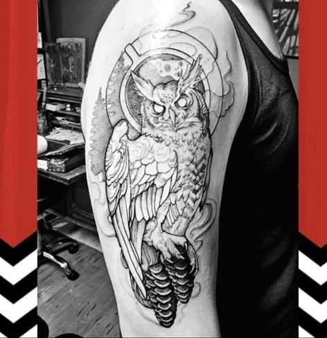 Zwart-witte tatoeage op de bovenarm van een persoon, met een gedetailleerde uil die met zijn poten rust op dennenappels. De achtergrond toont een kop koffie van bovenaf gezien, met stijgende damppluimen en contouren van bomen eromheen. Aan weerszijden van de afbeelding zijn illustraties van de iconische Red Room uit de serie Twin Peaks, herkenbaar aan de rode gordijnen en zwart-wit zigzagpatroon. De tatoeage verwijst naar de symboliek en mystiek uit de serie.