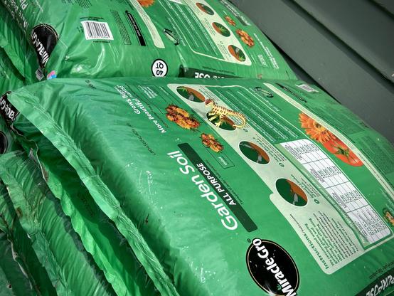 A green Dilophosaurus stands atop bright green bags of Miracle-Gro garden soil are stacked against a wall. The bags are labelled "All Purpose" and display images of flowers and planting instructions.