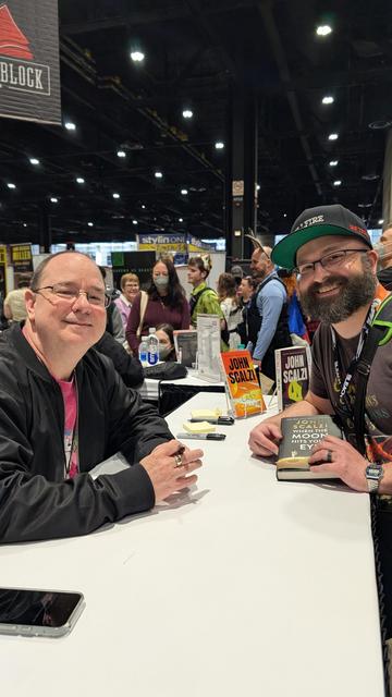 John Scalzi (left) and Derreck Mayer (right) posing at the former's signing table at C2E2 2025 in Chicago.