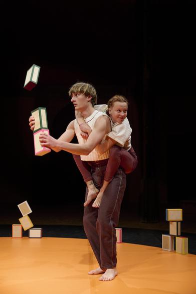 Un acrobate est debout au milieu de l'image. Il jongle avec 3 boîtes. Il a l'ai d'être concentré et regarde ses boites. L'autre acrobate est agrippée à lui dans son dos. Elle sourit avec un air espiègle.

Photo prise par L. d'Aboville