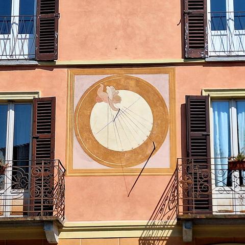 Close-up of a sundial painted on a building facade: a large circle with an angel in the top left, casting various painted rays toward the bottom right, where the numbers 12 to 19 are marked. There are two rods and a wire inbetween them, which casts a shadow in the direction of the rays.