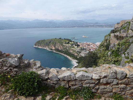 View shows part of dismantled wall in foreground from fortress of Palamidi, with dramatic drop below the town visible on a promontory jutting out into the bay. In the distance across the gulf are visible the mountains of the Peloponnese, Greece. 