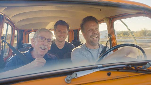 Das Bild zeigt eine fröhliche Szene aus dem Inneren eines alten orangefarbenen Autos während der Fahrt.

Drei Männer sitzen eng beieinander: Ein älterer Herr mit Brille und Schnurrbart sitzt vorne auf dem Beifahrersitz, ein jüngerer Mann steuert den Wagen, beide lachen herzlich. In der Mitte, auf dem Rücksitz, sitzt ein weiterer Mann, der ebenfalls breit lächelt. Die Sonne scheint durch die Fenster, das Licht ist warm und weich – es ist ein sonniger Tag, vermutlich in den Nachmittagsstunden.

Durch die Windschutzscheibe und die Seitenscheiben sieht man eine vorbeiziehende Landschaft mit Bäumen und einer Leitplanke – sie fahren offenbar auf einer Landstraße oder Autobahn. Die Stimmung ist ausgelassen, freundschaftlich und voller Leichtigkeit – ein Moment voller gemeinsamer Freude und Verbundenheit auf einer besonderen Reise.