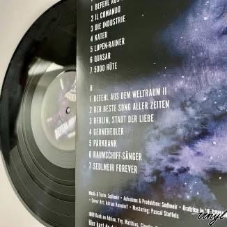 Abbildung einer Vinyl-Schallplatte mit einem Hintergrund mit Galaxienmotiven. Auf der Rückseite des Covers sind die Titel in deutscher Sprache aufgeführt, darunter "Befehl aus dem Weltraum II", "Der beste Song aller Zeiten" und "Berlin, Stadt der Liebe".
