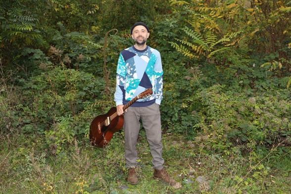 Der Singer-Songwriter Boo Hoo steht vor Pflanzen. Er trägt eine Pullover und in der Hand eine Gitarre.