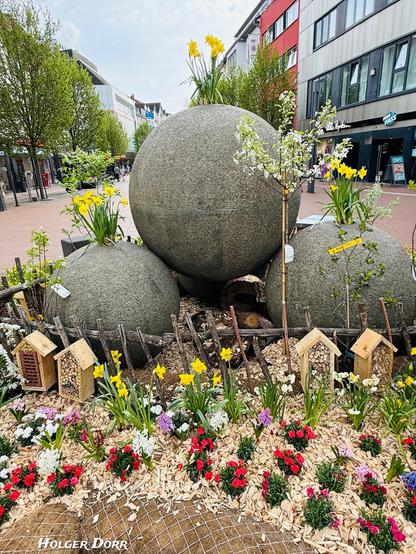 Nahaufnahme des österlich dekorierten Kugelbrunnens. Die großen Steinkugeln sind von bunten Frühlingsblumen, kleinen Insektenhotels und jungen Blütenbäumen umgeben. Ein harmonisches Zusammenspiel von Natur und urbanem Raum.