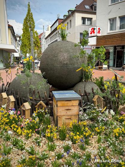 Der Kugelbrunnen mit zentral platzierter Bienenbeute, umgeben von gelben Narzissen, blühenden Sträuchern und kleinen Insektenhäusern. Die Szene wirkt wie ein kleines Frühlingsparadies mitten in der Stadt.
