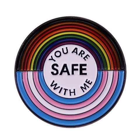 A circular badge with the upper semi-circle in Progress Pride rainbow colours and the lower semi-circle in Trans Flag colours, with the centre reading " You are SAFE with me"