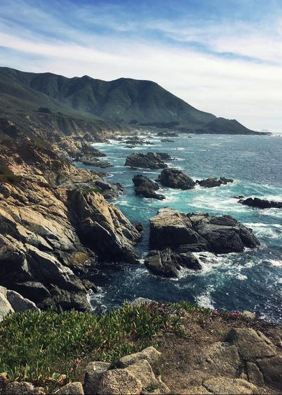 A cellphone photo of a Pacific Coast rocky shoreline with mountains to the left and ocean to the right. The sky is blue with many high, wispy clouds.
