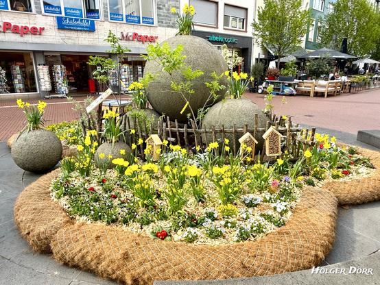 Der Kugelbrunnen in Gießen, festlich geschmückt als Osterbrunnen. Umgeben von bunten Frühlingsblumen, kleinen Insektenhotels und einer Bienenbeute, erstrahlt das Stadtwahrzeichen inmitten der Fußgängerzone in voller Blütenpracht. Jutematten umrahmen das Beet, während Passanten den farbenfrohen Anblick genießen.