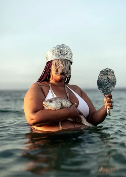 A corpulent woman with a headdress covering her face is holding a fish and a mirror while immersed in water.