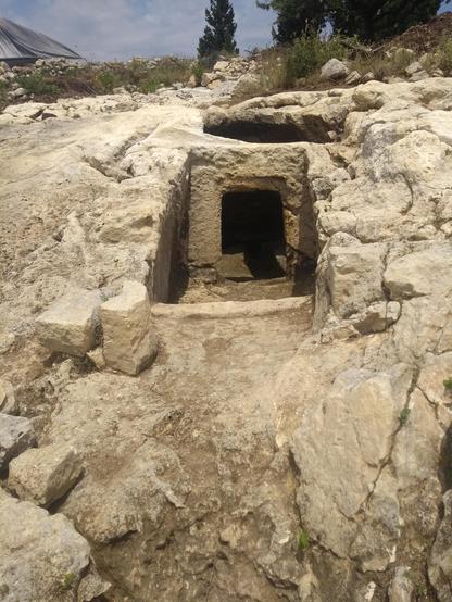 View of entrance to chamber tomb, set into rocky hillside with rectangular stone door frame.