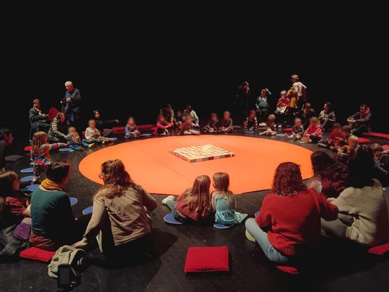 Photo d'un espace scénique orange en forme de cercle. Les gens sont assis par terre, en rond, tout autour de l'espace scénique. Au milieu du cercle orange, sont disposés 48 boîtes à cigare.