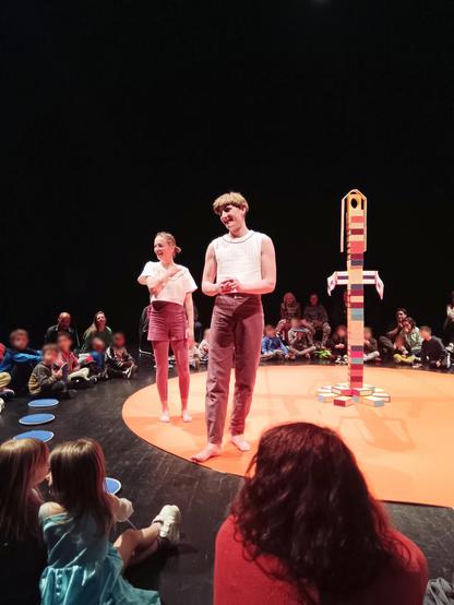 Les deux artistes acrobates sont au bord de l'espace scénique rond et orange. Ils ont l'air de discuter avec des personnes de l'audience. On peut voir au centre de l'espace scénique, à droite de l'image, une tour faîte de boîtes à cigares. 