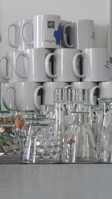 Ordentlich gestapelte Kaffeebecher in weiß, davor eine Sammlung von Trinkgläsern. Alle stehen kopf.