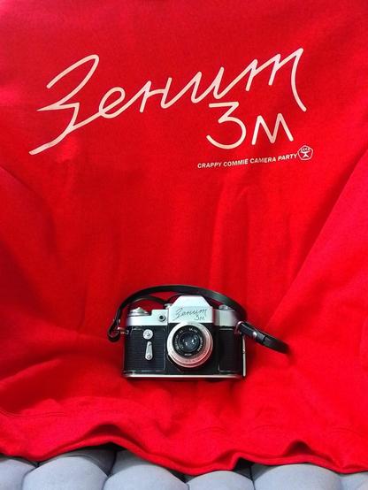 A red college shirt with a text "Zenit 3M" written ib cyrillic, and a corresponding Zenit 3M '64 camera lying on the shirt