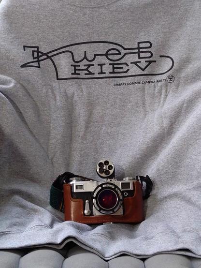 A grey college shirt with a text "Kiev" written in both cyrillic and latin characters, a Kiev 4A '61 rangefinder camera lying on top of it.
