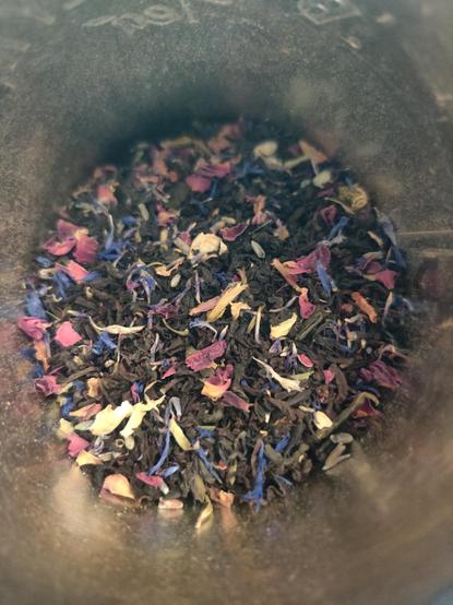 The camera is pointed into a tin of loose leaf tea, featuring black curled tea leaves and colorful floral inclusions.