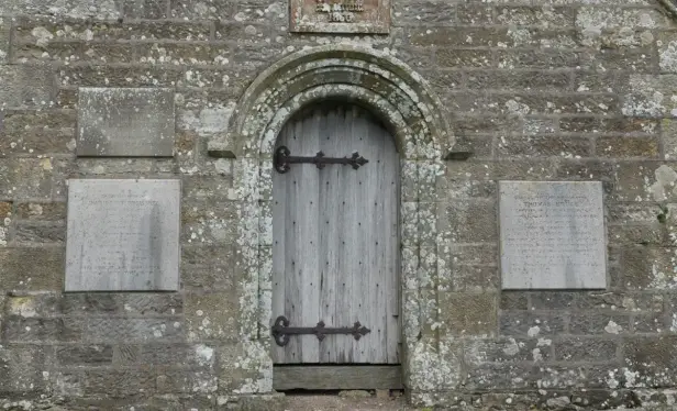 You see a grey old stone wall with lichens. In the middle a gray wooden door.
