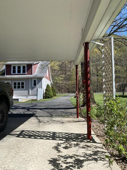 View from under a white carport ceiling looking across the late afternoon shadows on the cement at the gray, red and white farmhouse, with the spindly greening trees behind it, marching up the hillside from the creek across the little road