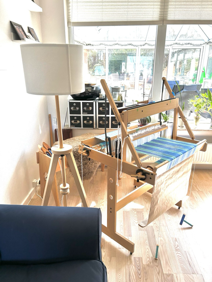A hand loom standing at an oblique angle in a corner of a room, beside a floor lamp and in front of a sunny window