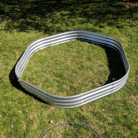 A photo showing a 10 foot by 4 foot raised bed assembled in an alternate method to create a hexagon shape. It is alternating a curved panel and a straight panel to complete the circumference. Normally it would be three curved panels on each end and 3 straight pieces per side. I don't think this shape will need any staking or support. It will make it difficult to get to the middle but I might put a large barrel container there that will have marigolds in it. I didn't measure it but it should be about 7 feet across the flats. 2.2 meters.