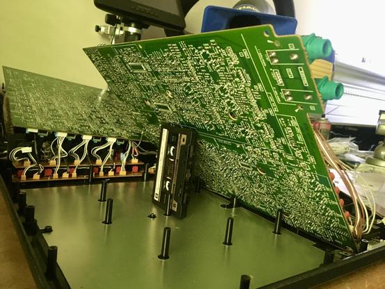 A view from underneath the R/P PCB of the Marantz PMD 740 multitrack cassette recorder. The PCB, which is bright green, is being partially propped up by a black Maxell cassette tape standing on its side.