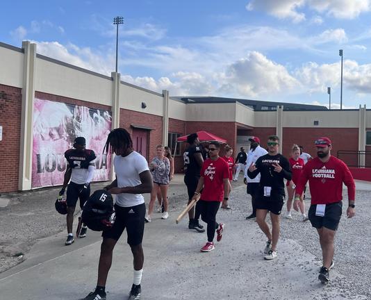 A group of football players walk back from practice in front of a one story building. Blue sky. Clouds. Old faded mural on the building wall behind them that you cannot quite make out. 