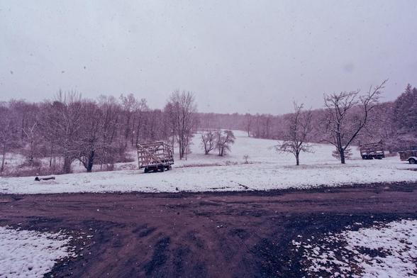 A snow covered hill with a melted dirt driveway forming a T-shape facing the camera and past that two hay wagons and a few small trees in a flat section before a field works its way up a hill framed by winter phase trees