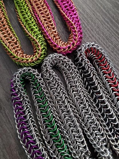 on a gray wood desktop lays five fidget toys which have an elongated mobius shape. they are made with various colors and materials. on top are two fidgets in anodized aluminum in pink, green, and copper. bottom three include an all stainless steel version paired on either side with stainless steel paired with stripes of anodized aluminum. 