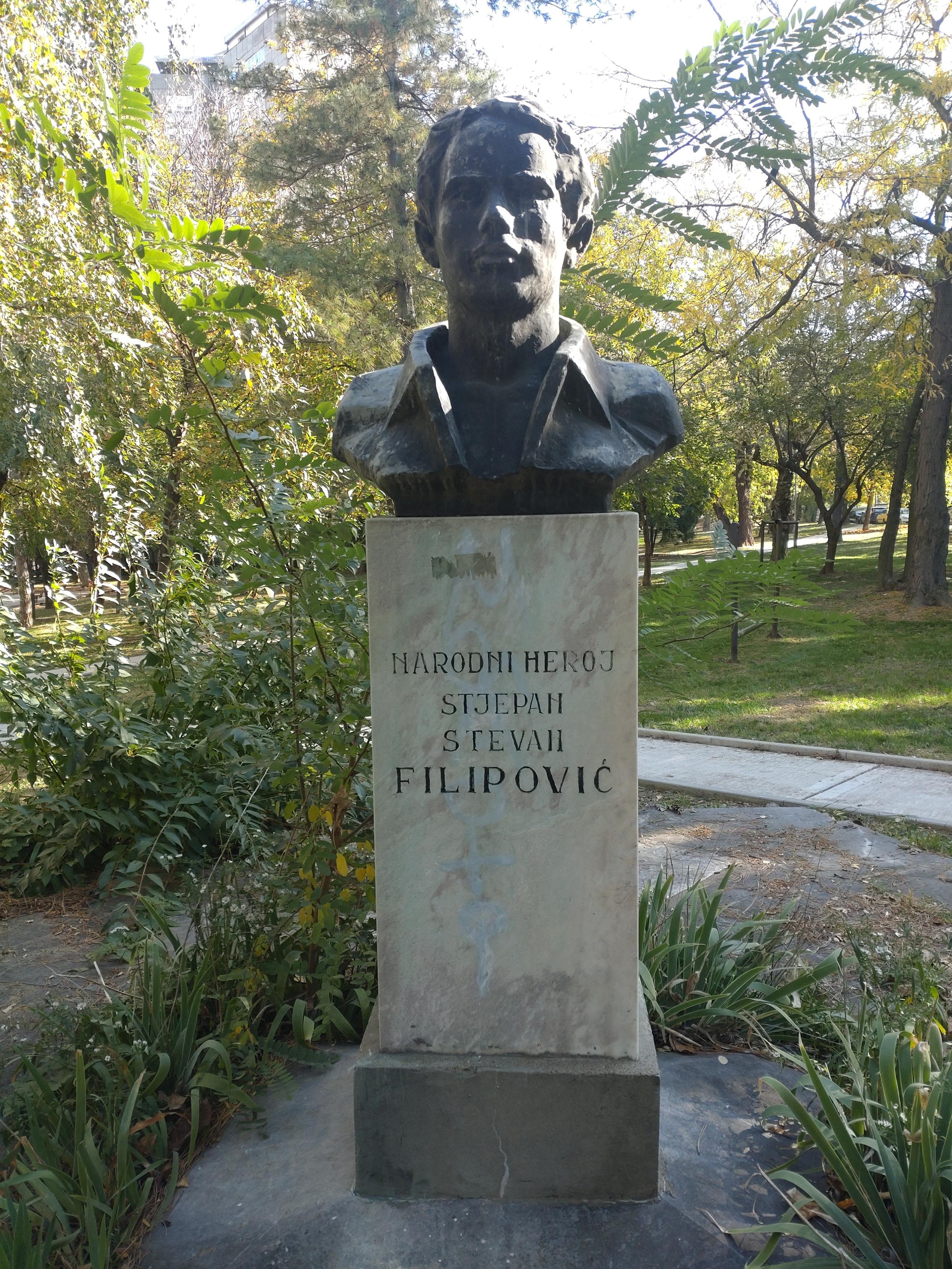An einem sonnigen Tag in einer Grünanlage mit schmalen befestigen Wegen steht im Zentrum des Bildes das Denkmal von Stjepan Filipovic. Auf einem Sockel ist lediglich sein Kopf sowie Jackenkragen platziert. An einem sonnigen Tag in einer Grünanlage mit schmalen befestigen Wegen steht im Zentrum des Bildes das Denkmal von Stjepan Filipovic. Auf einem Sockel ist lediglich sein Kopf sowie Jackenkragen platziert.