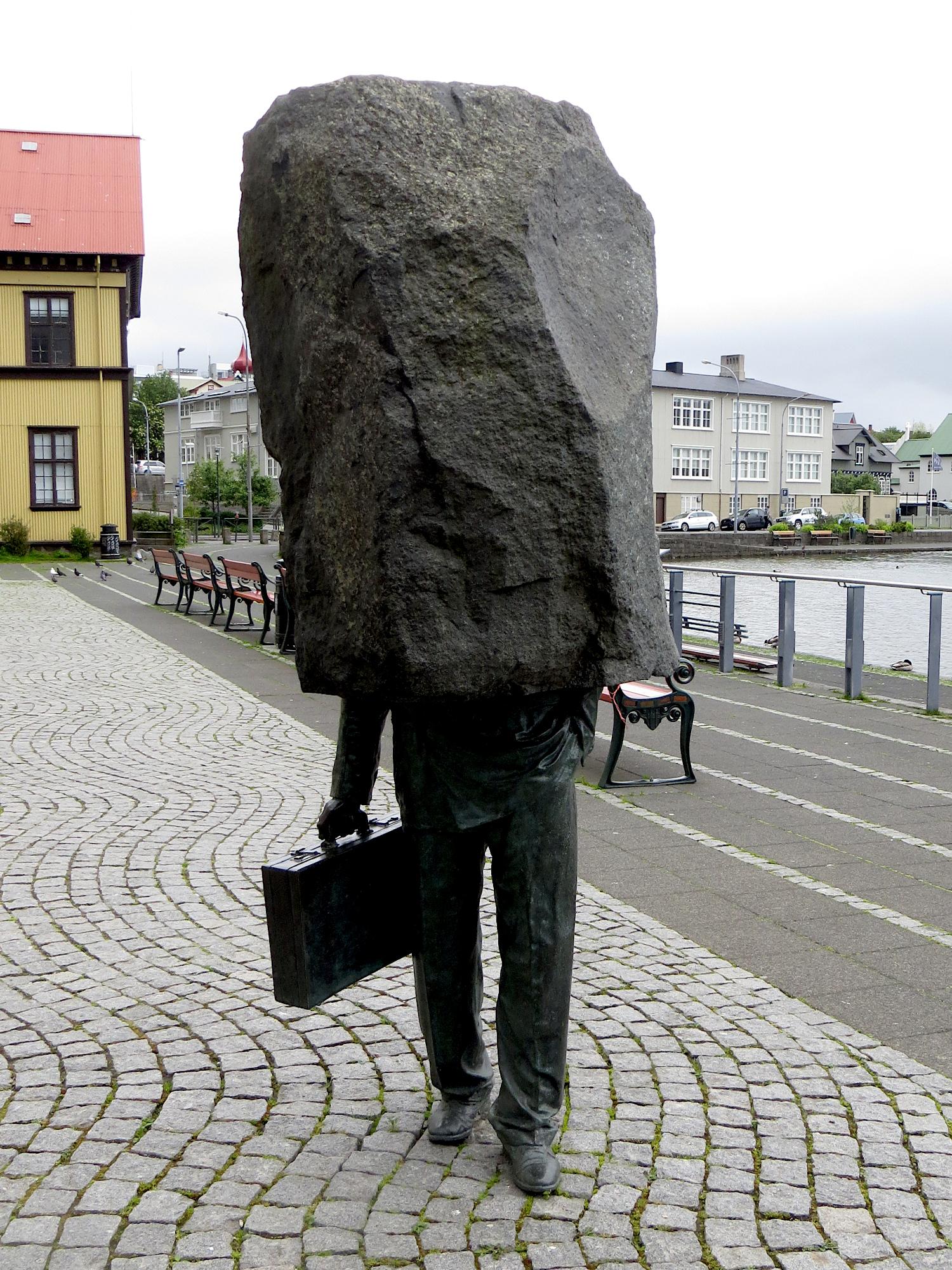 Das Bild zeigt das Denkmal für den 'Unbekannten Bürokraten' (orginal: The Unknown Bureaucrat) in Reyjkavik, Island. Das Denkmal zeigt eine Person im Anzug, in der rechten Hand trägt sie einen Aktenkoffer. Aber von etwa der Hüfte an Aufwärts ist nur ein grober Felsblock zu sehen. Als hätte der Künstler in der Mitte die Arbeit eingestellt. Das Bild zeigt das Denkmal für den 'Unbekannten Bürokraten' (orginal: The Unknown Bureaucrat) in Reyjkavik, Island. Das Denkmal zeigt eine Person im Anzug, in der rechten Hand trägt sie einen Aktenkoffer. Aber von etwa der Hüfte an Aufwärts ist nur ein grober Felsblock zu sehen. Als hätte der Künstler in der Mitte die Arbeit eingestellt.