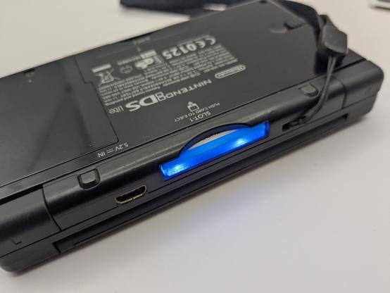 A black ds lite closed face down exposing the cart port showing a blue ds lite cart lite up blue