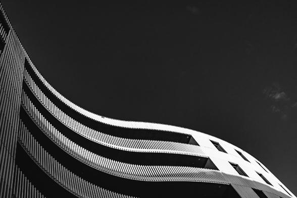 Black and white photo of a modern architectural building with wavy, horizontal lines and a rhythmic pattern of vertical slats. The curves of the structure contrast sharply against a clear, dark sky, giving the image a dynamic and minimalist feel.

