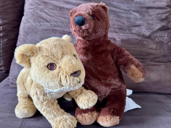 A stuffed lion and a stuffed brown bear are sitting together on a brown couch. The lion is light tan with orange eyes, while the bear is dark brown with a black nose.